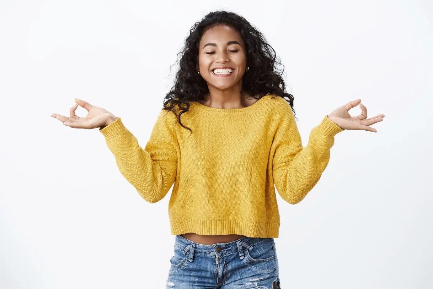 Opgewekt, zorgeloos, vrolijk meisje met krullend haar sluit haar ogen en ademt frisse lucht in, glimlacht opgelucht en vrij, mediteert met zijwaarts gespreide handen in mudra/zen-gebaren, witte muur.